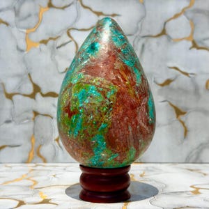 Op de afbeelding: Een gepolijst, eivormig stenen beeldhouwwerk met een levendige mix van dieproestrode, helder turquoise en mosgroene mineraalpatronen. Het gladde, druppelvormige object is bevestigd op een kleine, ronde, donkerbruine houten voet.