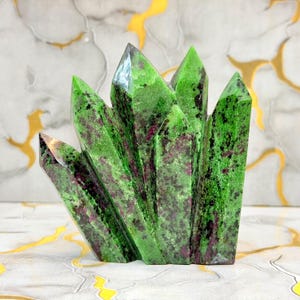 Può includere: Un gruppo di cristalli naturali di zoisite caratterizzato da un colore di base verde brillante con inclusioni magenta intenso e viola scuro. L'esemplare è composto da molteplici formazioni cristalline appuntite e verticali fuse alla base.