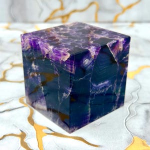 Puede incluir: Un cubo pulido tallado en piedra de fluorita natural, que muestra tonos profundos de púrpura, índigo y azul translúcido con fracturas cristalinas internas visibles y bandas naturales en toda la estructura geométrica.