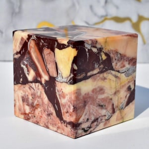 May include: A square, decorative stone block with a polished surface. The cube features a mix of colors, including deep burgundy, pale yellow, and light pink, creating a marbled effect. The stone has a natural, organic appearance.
