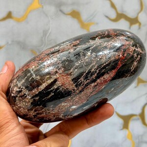 Puede incluir: Una piedra pulida de forma ovalada que presenta un color base negro oscuro con vetas irregulares y parches prominentes de inclusiones minerales de color rojo intenso y plata metálica en toda su superficie lisa y reflectante.