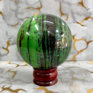 Puede incluir: Una esfera de piedra preciosa pulida, predominantemente verde con vetas negras y rojas. La esfera se exhibe sobre un pequeño soporte redondo de madera rojo oscuro. El fondo presenta un patrón similar al mármol con detalles dorados.