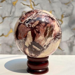 Può includere: Una sfera di pietra lucida, appoggiata su un supporto di legno scuro. La sfera presenta un mix di colori crema, bordeaux e nero, con intricati motivi di venature. Il supporto è di colore marrone scuro.