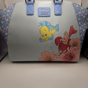 May include: A light blue handbag featuring illustrations of Flounder and Sebastian from Disney's The Little Mermaid. The bag has light blue handles and a Disney Loungefly logo. The sides of the bag have a patterned design with stars and sea creatures.
