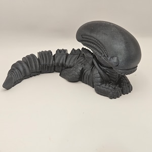 May include: A black, articulated Xenomorph figure, a creature from the Alien film franchise. The figure is lying down, with a detailed head and segmented body. The figure is made of a matte black material.