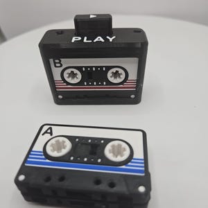 May include: Two black cassette tape-shaped objects. One has the word "PLAY" on top and a cassette tape design. The other is a smaller cassette tape with a blue stripe design. Both have white and red accents.
