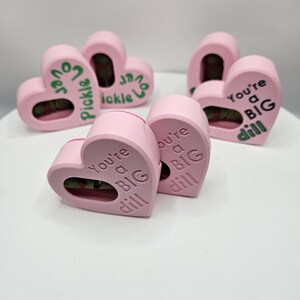 May include: Pink heart-shaped containers with the phrases "Pickle Lover" and "You're a BIG dill" printed on them. The containers have a small window to view the contents. The objects are arranged on a white surface.