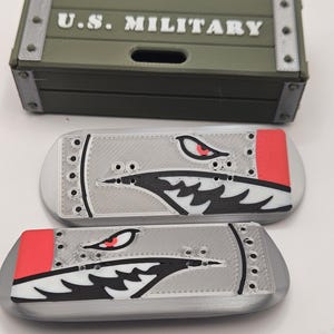 May include: A green rectangular box with the text "U.S. MILITARY" in white, above two silver-colored, oval-shaped objects. Each oval has a shark-like design with red, black, and white accents.