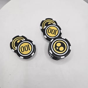 May include: A collection of circular gaming chips in black, white, and gold. Some chips feature the number 100 printed in gold, while others display a gold skull icon. The chips have a textured edge with alternating black and white rectangular segments.