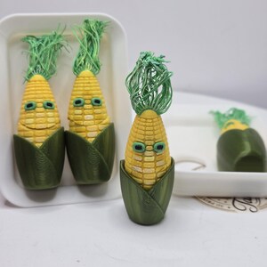 May include: Four small, yellow corn-shaped figurines with green husks and green fibrous tops. Each corn figure features a face with two dark green eyes and a subtle mouth. The figures are arranged in white rectangular trays on a white surface.