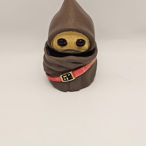 May include: A brown figurine of a hooded character with a gold face and red belt. The figure has a pointed hood and a wrapped body. The eyes are dark and the belt has a gold buckle. The figurine is on a white background.