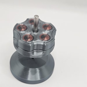 May include: A gray, 3D-printed object with a cylindrical base and a top resembling a revolver cylinder. The top has six circular indentations with copper-colored details. A silver rod extends from the center.