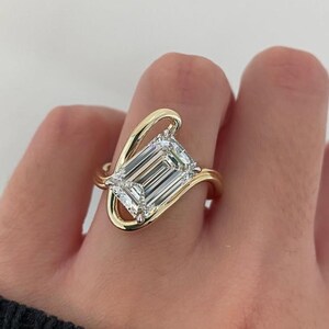 May include: A gold ring featuring a large, rectangular emerald-cut clear gemstone set within a smooth, asymmetrical, flowing gold band that curves around the stone, creating an open, continuous loop design that encircles the center stone.