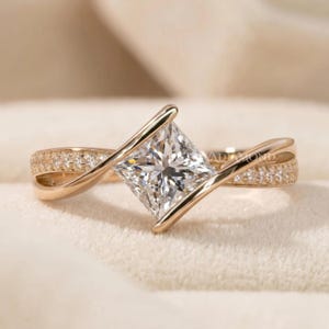 Könnte beinhalten: Ein Ring aus Roségold mit einem zentralen Diamanten im Prinzessschliff, der von zwei diagonalen Bändern gehalten wird. Die Ringschiene ist in zwei sich kreuzende Abschnitte unterteilt, die jeweils mit einer Reihe kleiner Pavé-Diamanten besetzt sind.
