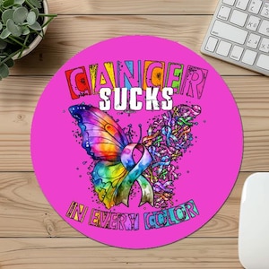Puede incluir: Una alfombrilla de ratón redonda y rosa con un diseño de mariposa colorido y el texto "CANCER SUCKS IN EVERY COLOR". La mariposa tiene alas de arcoíris y una cinta. La alfombrilla está sobre un escritorio de madera junto a un teclado y un ratón.