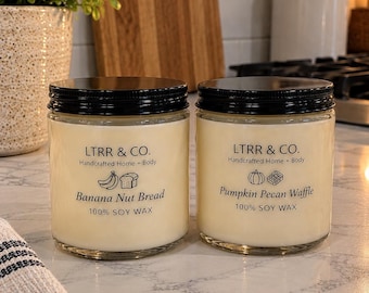 May include: Two glass jar candles with black lids and cream-colored soy wax. The left label reads LTRR & CO. Handcrafted Home + Body, Banana Nut Bread, 100% SOY WAX with banana and bread icons. The right label reads Pumpkin Pecan Waffle with icons.