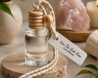 May include: A clear glass car diffuser bottle with a wooden cap and a white and gold braided cord. A rectangular white tag attached to the cord features the black printed text Love The Reiki Roam Car Diffuser. The bottle contains clear liquid.