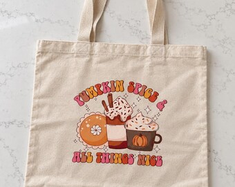 May include: A beige canvas tote bag featuring a graphic print of a pumpkin pie, a tall drink with whipped cream and cinnamon sticks, and a mug with a pumpkin design. The text reads PUMPKIN SPICE & ALL THINGS NICE in a retro, multi-colored font.