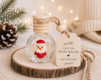 May include: A clear glass car diffuser bottle with a wooden cap and a small Santa Claus figurine inside. A beige rectangular tag is attached with a cord, featuring the text LOVE THE REIKI ROOM and CAR DIFFUSER with small heart and leaf illustrations.
