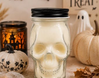 May include: A clear glass jar shaped like a human skull, filled with off-white wax. The jar features a black metal screw-top lid. The skull design includes molded eye sockets, a nasal cavity, and teeth, creating a distinct anatomical appearance.