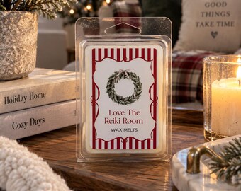 May include: A clear plastic clamshell container holding wax melts with a white label featuring a green pine wreath illustration, red and white vertical stripes, and the text Love The Reiki Room WAX MELTS printed in a clear, dark red font.