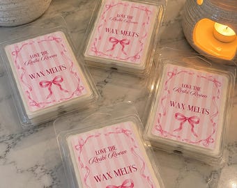 May include: Four rectangular clear plastic clamshell containers filled with wax melts. Each container features a white and pink striped label with a pink ribbon illustration and the text: LOVE THE Reiki Room WAX MELTS in pink lettering.