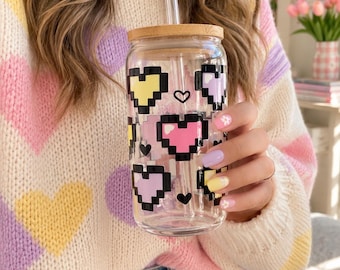 May include: A clear glass tumbler with a bamboo lid and a clear glass straw. The exterior of the glass features a repeating pattern of pixelated hearts in pastel shades of yellow, pink, and lavender, each outlined in a bold black border.