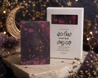 May include: A rectangular soap bar with a dark purple and magenta marbled surface featuring gold flecks. It stands next to a white cardboard box with a window, labeled LOVE THE REIKI ROOM SOAP BAR, Traditional Cold Process Soap, Vegan, Cruelty Free, Phthalate Free Fragrance, Made in the USA.