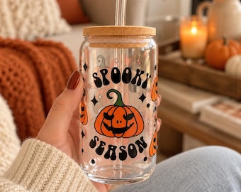 May include: A clear glass tumbler with a bamboo lid and a glass straw. The exterior features a black graphic print that reads "SPOOKY SEASON" in a wavy font, accompanied by orange pumpkin illustrations and small black star accents.