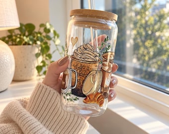 May include: A clear glass tumbler with a bamboo lid and a glass straw, featuring a colorful graphic print of various coffee drinks, a croissant, and small green leaves. The design includes a tall iced coffee with whipped cream and a heart.