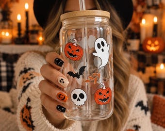 May include: A clear glass tumbler with a bamboo lid and a glass straw, featuring printed Halloween-themed graphics including orange jack-o'-lanterns, a white ghost, a black bat, a white skull, and small orange leaves scattered across the surface.