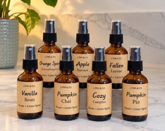 May include: Seven amber glass spray bottles with black pump tops, each featuring a tan label with the brand name LTRR & CO. and text identifying scents including Vanilla Bean, Orange Spice, Apple Harvest, Pumpkin Chai, Cozy Campfire, Fallen Leaves, and Pumpkin Pie.