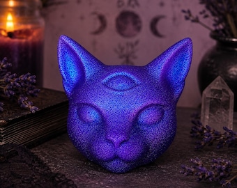 May include: A three-eyed cat head sculpture with a vibrant, iridescent purple and blue finish. The surface has a fine, textured, glittery appearance. The cat has closed eyes and pointed ears, with a third eye centered on the forehead.