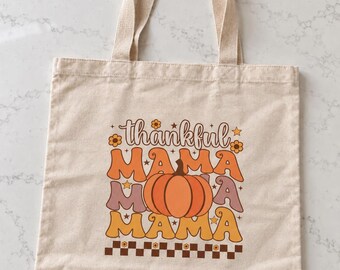 May include: A beige canvas tote bag featuring a graphic print with the text "thankful MAMA MAMA MAMA" in orange, yellow, and muted purple wavy lettering. A central orange pumpkin illustration and a brown checkered border complete the design.