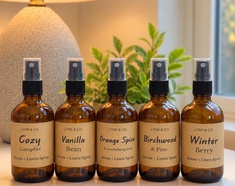 May include: Five amber glass spray bottles with black pump nozzles, each featuring a tan label with the text LTRR & CO. and scent names: Cozy Campfire, Vanilla Bean, Orange Spice & everything nice, Birchwood & Pine, and Winter Berry Room + Linen Spray.
