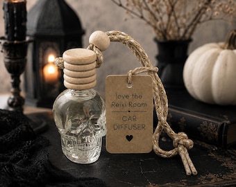 May include: A clear glass skull-shaped car diffuser bottle with a light wood cap and a braided rope hanger. A rectangular kraft paper tag is attached, featuring the printed text: love the Reiki Room, CAR DIFFUSER, and a small heart symbol.