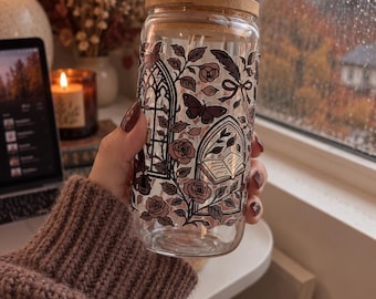 May include: A clear glass tumbler with a bamboo lid and a glass straw, featuring a printed design of gothic arched windows, open books, blooming roses, butterflies, and feathers in shades of muted pink, brown, and black ink outlines.