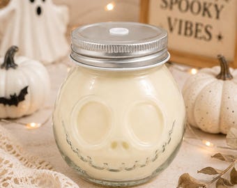 May include: A clear glass jar with a rounded shape and a skull-like face embossed on the front, filled with a creamy off-white substance. The jar features a silver-colored metal lid with a small white circular center piece and a straw hole.