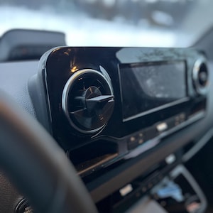 Op de afbeelding: Een close-up van een autodashboard met een glanzend zwarte afwerking. Het dashboard bevat ventilatieopeningen met zilveren afwerking, een digitaal beeldscherm en diverse bedieningsknoppen. Het stuurwiel is zichtbaar op de voorgrond.