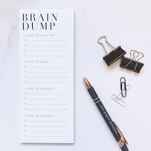 May include: A white notepad with the words "BRAIN DUMP" and sections for notes, along with a black pen, a black and gold pen, binder clips, and a paper clip. The black and gold pen has the words "art paper scissors" printed on it.