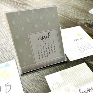 May include: A gray desktop calendar with a raindrop pattern and the word "april" written in brown script. The calendar shows the month of April with the days of the week labeled S, M, T, W, T, F, S. The numbers 1 through 30 are listed on the calendar.