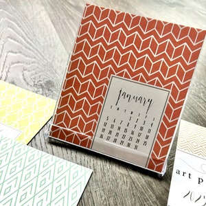 May include: A desktop calendar with a geometric pattern in shades of orange and white. The calendar is for January and shows the days of the week and dates. The calendar is on a wooden surface.