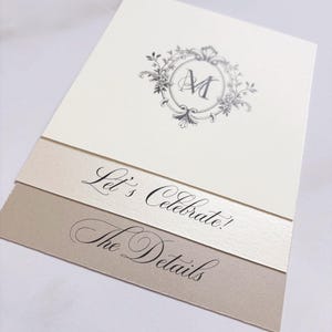 May include: Ivory and beige wedding invitation suite with a floral crest and the letter "M". The text "Let's Celebrate!" and "The Details" are displayed in elegant script. A gold brad secures the layers.