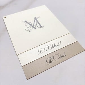 May include: Ivory-colored invitation card with a silver monogram and the words "Let's Celebrate!" and "The Details" in elegant script. The card has a layered design with a gold accent. A small gold stud is in the upper left corner.
