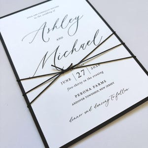 May include: A formal wedding invitation with black text on a white background, featuring the names "Ashley" and "Michael" in elegant script. The invitation is bound with black twine and has a black border. The text includes the date, time, and location.