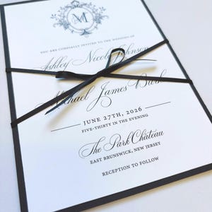 May include: Wedding invitation with a black border and a black ribbon. The invitation is white with black text and a blue floral crest with the letter "M". The text includes the names Ashley Nicole Johnson and Michael James Burke, and the date June 27th, 2026.