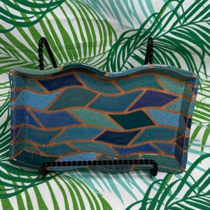 May include: A rectangular ceramic tray with wavy edges, featuring a mosaic-like pattern of blue, teal, and dark purple geometric shapes outlined in copper. The surface has a speckled texture and is displayed on a black wire stand.