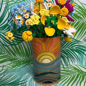 May include: A cylindrical ceramic vase featuring a painted sunset and ocean landscape in shades of orange, yellow, and blue, filled with a colorful bouquet of plastic building block flowers in yellow, white, blue, and purple hues.
