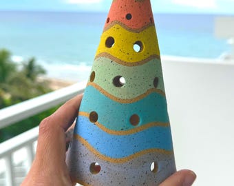 May include: A conical ceramic sculpture shaped like a tree with a yellow star topper. The surface features horizontal wavy bands in shades of red, orange, yellow, green, teal, blue, and purple, all speckled with black dots and circular cutouts.