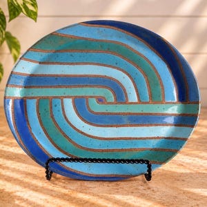 May include: A round ceramic plate featuring a swirling pattern of concentric stripes in shades of dark blue, light blue, and teal. The design is separated by thin, exposed brown clay lines, and the surface has a speckled texture.